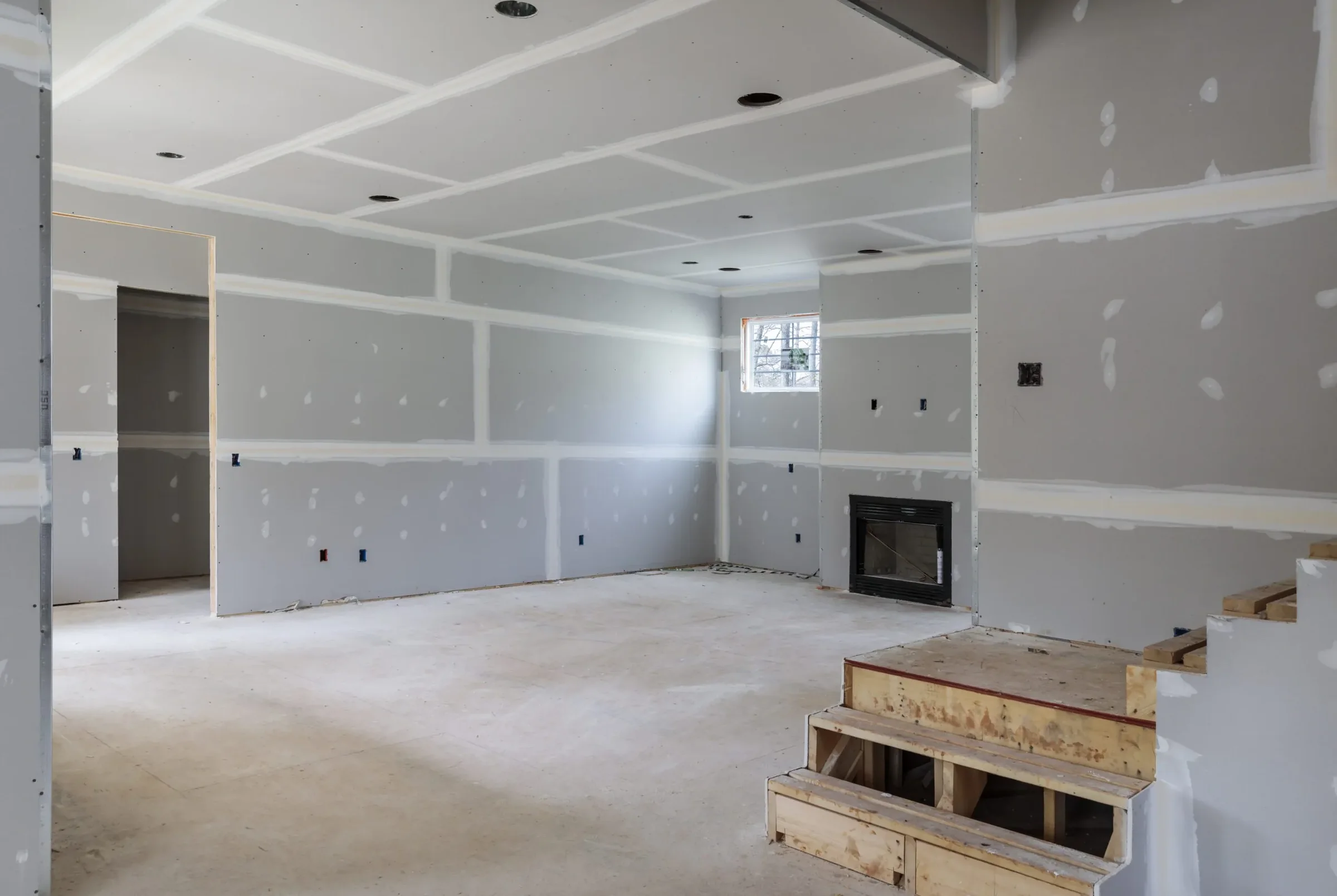 Room with drywall hung and taped, showing a fireplace opening, recessed cans and an unfinished floor
